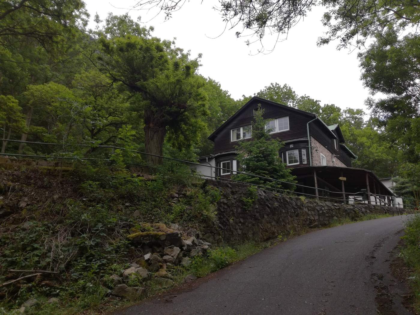 Dřevěná Chata pod Bořeněm nad úzkou asfaltovou silničkou, kolem stromy a kamenná zeď