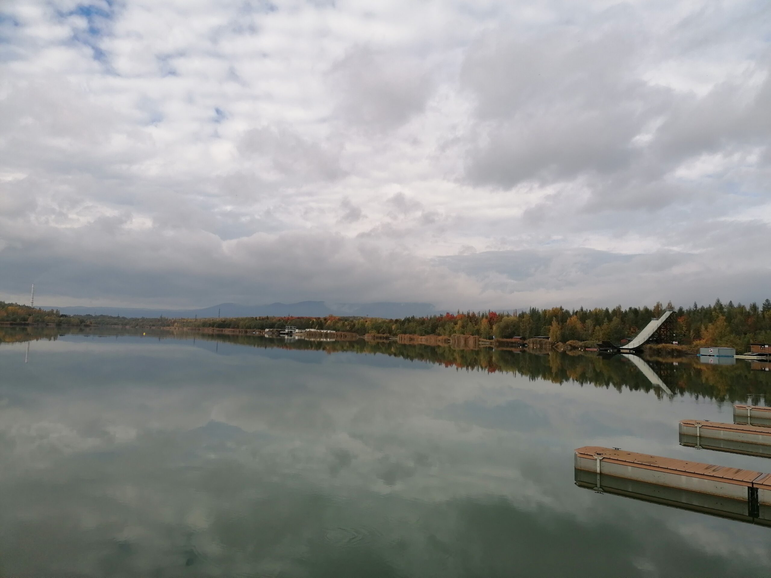 Klidná hladina jezera Matylda s odrazem mraků a podzimní krajiny. Skokanský můstek a mola. Rekreace a vodní sporty v přírodě.